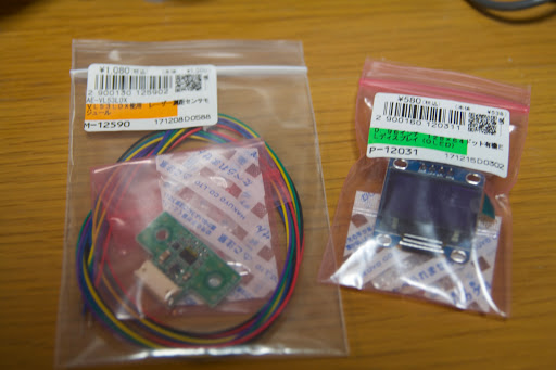 机の上に置かれた秋月電子の袋入り部品。左はVL53L0Xレーザー測距モジュールとカラー配線、右は小型ディスプレイモジュール